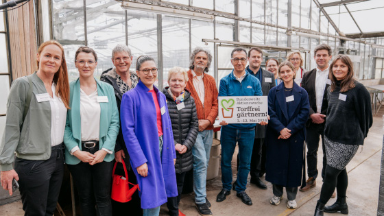 Gruppenbild in einem Gewächshaus Bundesminister Özdemir hält ein Schild mit dem Schriftzut "Bundesweite Aktionswoche Torffrei gärtnern! 3. - 12 Mai 2024" in den Händen