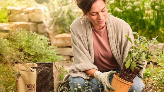 Eine Frau kniet in einem Garten und pflanzt eine Pflanze ein, neben ihr steht ein Sack Blumenerde